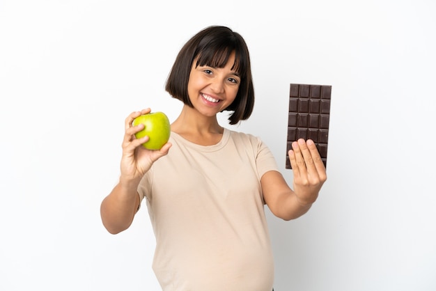 Jovem mestiça grávida isolada no fundo branco pegando um tablete de chocolate em uma das mãos e uma maçã na outra