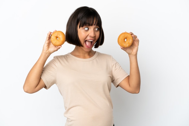 Jovem mestiça grávida isolada no fundo branco segurando rosquinhas e surpresa