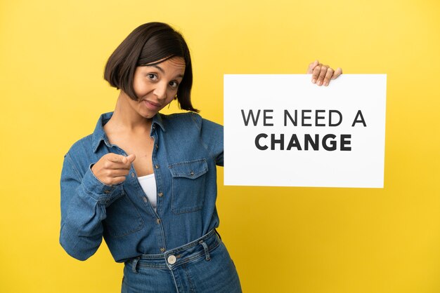 Jovem mestiça isolada em um fundo amarelo segurando um cartaz com o texto Precisamos de uma mudança e apontando para a frente