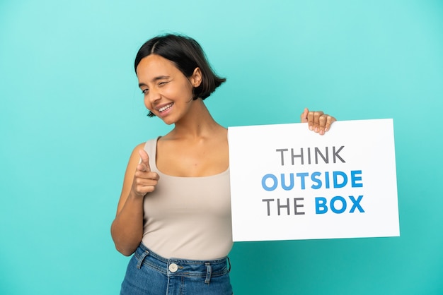 Jovem mestiça isolada em um fundo azul segurando um cartaz com o texto Pense fora da caixa e apontando para a frente