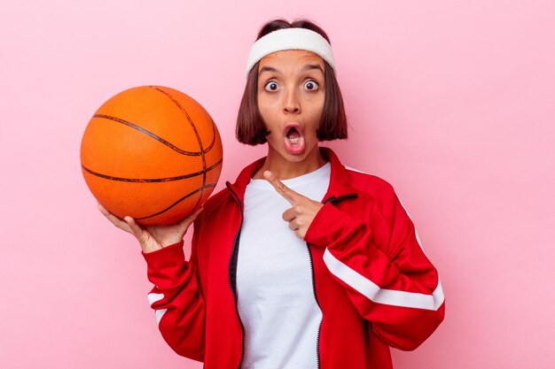 Jovem mestiça jogando basquete isolada em uma parede rosa apontando para o lado