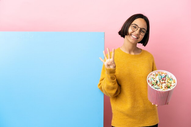 Jovem mestiça segurando pipocas com um grande banner sobre um fundo isolado feliz e contando quatro com os dedos