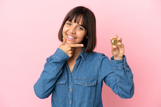 Foto jovem mestiça segurando um fundo isolado de bitcoin, feliz e sorridente