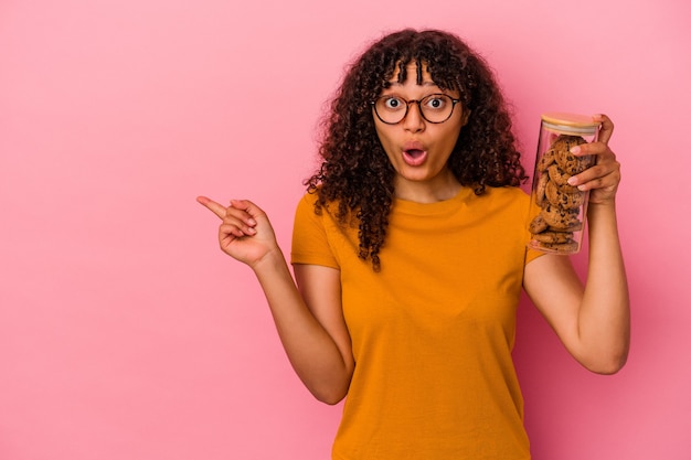 Jovem mestiça segurando um pote de biscoitos isolado na parede rosa apontando para o lado