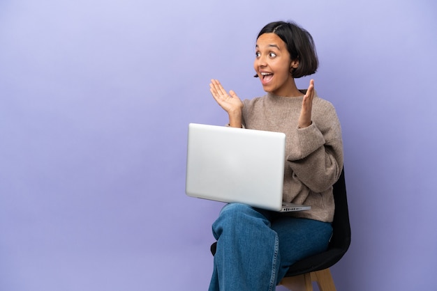 Jovem mestiça sentada em uma cadeira com laptop isolada com expressão facial surpresa