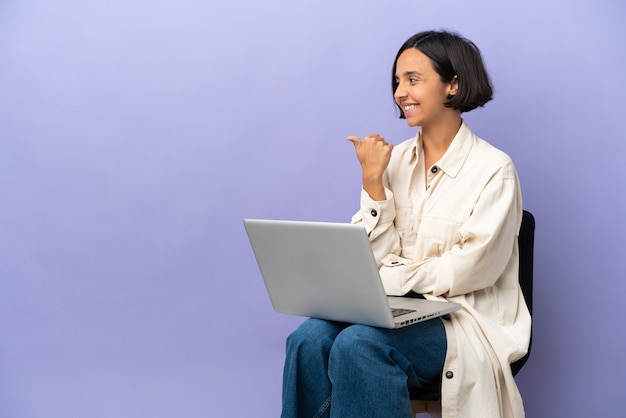 Jovem mestiça sentada em uma cadeira com laptop isolado em um fundo roxo apontando para o lado para apresentar um produto