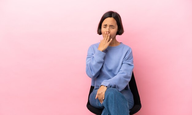 Jovem mestiça sentada em uma cadeira isolada em um fundo rosa, bocejando e cobrindo a boca aberta com a mão