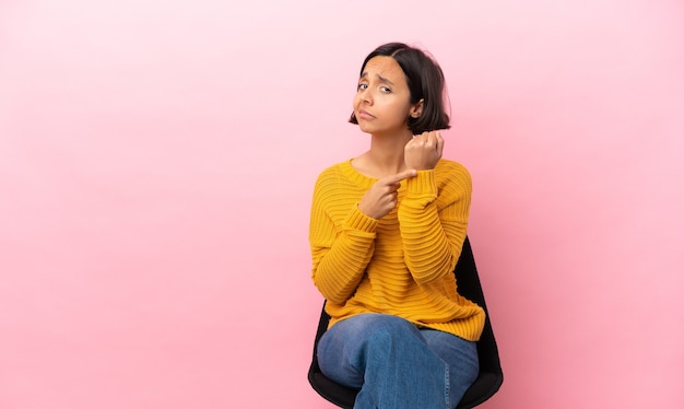 Jovem mestiça sentada em uma cadeira isolada em um fundo rosa fazendo o gesto de se atrasar