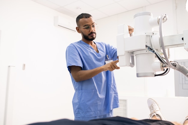 Jovem mestiço barbudo em uniforme azul usando novo equipamento médico em clínicas enquanto atendia um dos pacientes