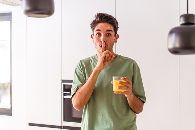 Jovem mestiço bebendo suco de laranja em sua cozinha, mantendo um segredo ou pedindo silêncio.