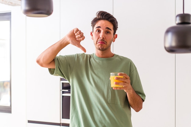 Jovem mestiço bebendo suco de laranja em sua cozinha, mostrando um gesto de antipatia, polegares para baixo. conceito de desacordo.
