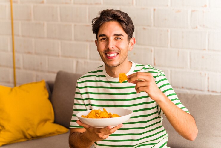 Foto jovem mestiço comendo batatinhas sentado no sofá