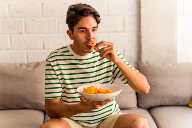 Jovem mestiço comendo batatinhas sentado no sofá