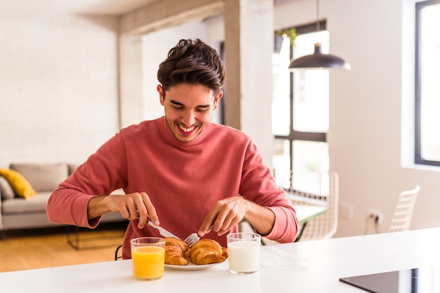 Jovem mestiço comendo croissant em uma cozinha pela manhã