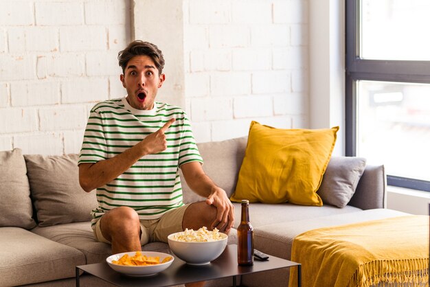 Jovem mestiço comendo pipoca sentado no sofá apontando para o lado