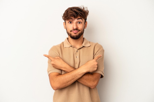 Jovem mestiço isolado em um fundo cinza aponta para os lados, está tentando escolher entre duas opções.