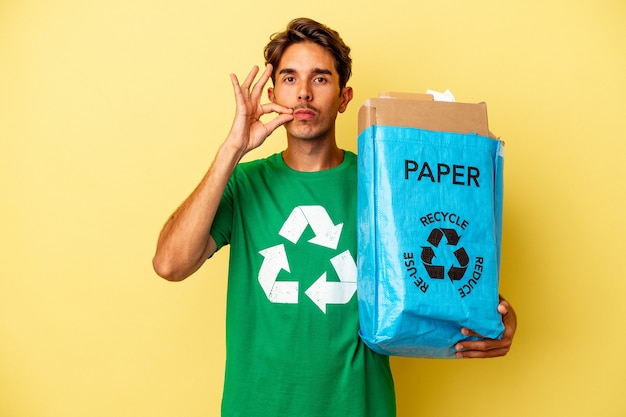 Jovem mestiço reciclando papel isolado em um fundo amarelo com os dedos nos lábios, mantendo um segredo.