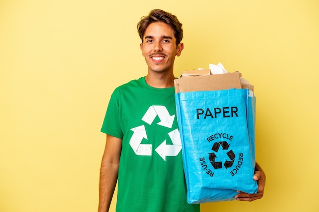 Jovem mestiço reciclando papel isolado em um fundo amarelo feliz, sorridente e alegre.
