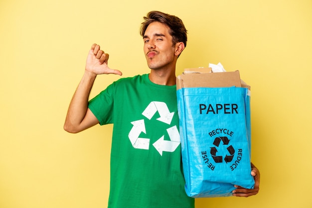 Jovem mestiço reciclando papel isolado em um fundo amarelo se sente orgulhoso e autoconfiante, exemplo a seguir.