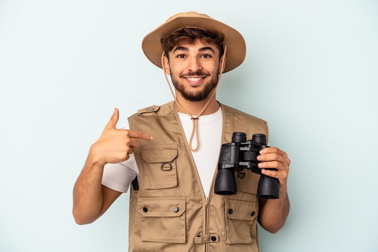 Foto jovem mestiço segurando binóculos isolados em um fundo azul pessoa apontando com a mão para um espaço de cópia de camisa, orgulhoso e confiante