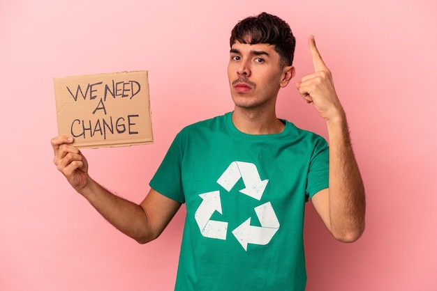 Jovem mestiço segurando um cartaz de "precisamos de uma mudança" isolado em um fundo rosa