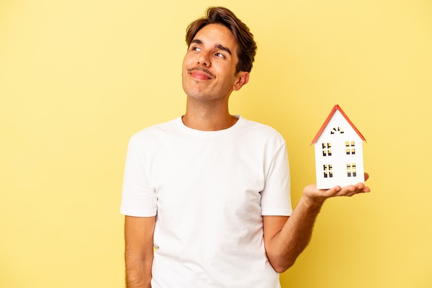 Jovem mestiço segurando uma casa de brinquedo isolada em um fundo amarelo, sonhando em alcançar objetivos e propósitos