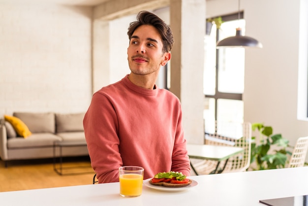 Jovem mestiço tomando café da manhã em sua cozinha parece de lado sorrindo, alegre e agradável.