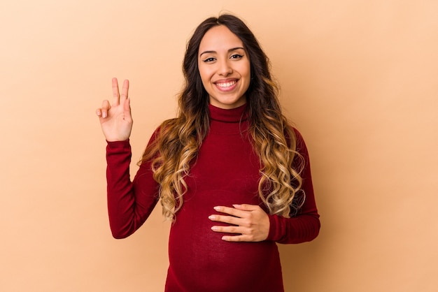 Jovem mexicana grávida isolada em um fundo bege, mostrando o sinal da vitória e sorrindo amplamente.