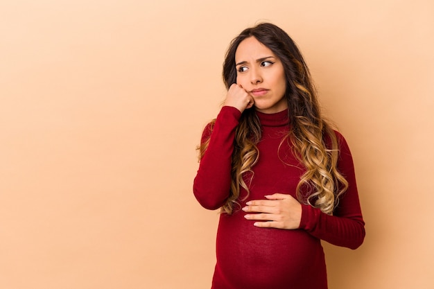 Jovem mexicana grávida isolada na parede bege que se sente triste e pensativa, olhando para o espaço da cópia.