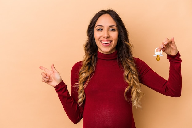 Jovem mexicana grávida segurando chupeta isolada em fundo bege, sorrindo e apontando de lado, mostrando algo no espaço em branco.