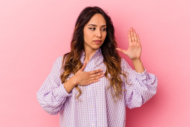 Jovem mexicana isolada em um fundo rosa, fazendo um juramento, colocando a mão no peito.