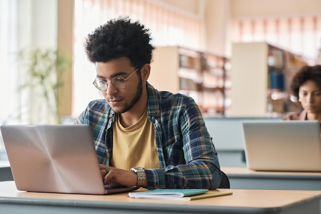 Jovem migrante sentado na mesa e estudando online com o laptop com outros alunos da classe