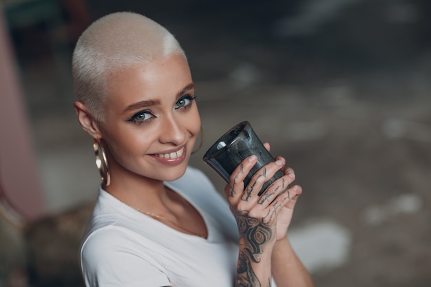 Jovem milenar com retrato de cabelo loiro curto sentado e sorrindo com uma xícara de chá ou uma bebida de café no sofá vintage