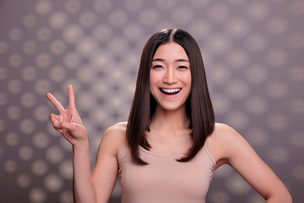 Jovem modelo asiático otimista levantando a mão e mostrando o símbolo da paz com o retrato de dois dedos. Mulher feliz sorridente posando com sinal de vitória, rindo e olhando para a câmera