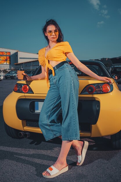 Jovem modelo com blusa azul jeans laranja e óculos escuros, ela está segurando um copo de café de papel encostado no carro amarelo no estacionamento