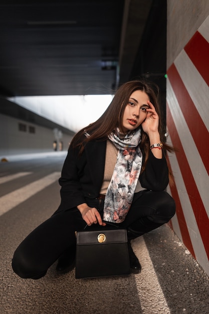 Jovem modelo elegante com roupas da moda com lenço vintage em botas com bolsa de couro preto da moda é relaxa no estacionamento perto do pilar listrado. Menina de moda elegante posando na cidade.