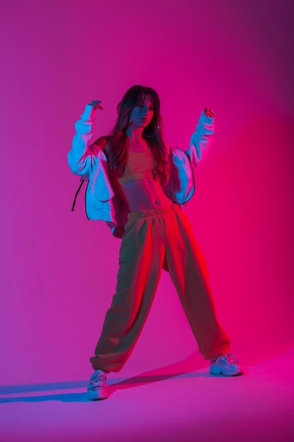 Jovem modelo elegante em roupas esportivas elegantes em sapatos posando no estúdio com cor ultravioleta multicolorida. Dançarina de linda garota dançando em uma sala com luzes de néon rosa brilhantes. Estilo.
