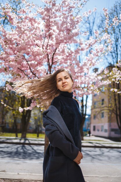 Jovem modelo esguia, com cabelos longos ondulados e, vestida com um casaco cinza, tênis, girando na rua. A flor da primavera, árvores, mulher, menina, ri e corre para se alegrar