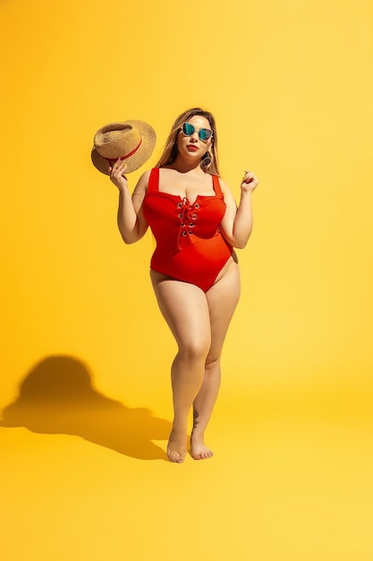 Jovem modelo feminino caucasiano plus size está se preparando para o resort de praia em fundo amarelo. Mulher de maiô vermelho, chapéu e óculos escuros. Conceito de verão, festa, corpo positivo, igualdade e descontração.