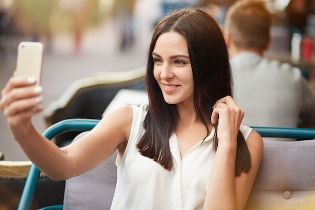 Jovem modelo feminino com cabelo liso escuro e sorriso gentil posa para fazer selfie, senta-se contra café de calçada ao ar livre, usa telefone inteligente moderno. Mulher européia encantada positiva no restaurante