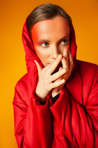 Jovem modelo feminino em um elegante casaco vermelho, segurando a mão no bolso e tocando o rosto com maquiagem criativa contra um fundo amarelo