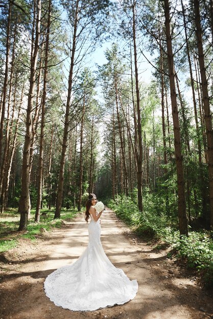 Jovem modelo mulher com vestido de noiva elegante de renda, mantendo um buquê e posando na floresta.