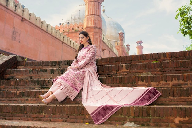 Foto jovem modelo vestindo vestido floral roxo sentada nas escadas mesquita badshahi no fundo