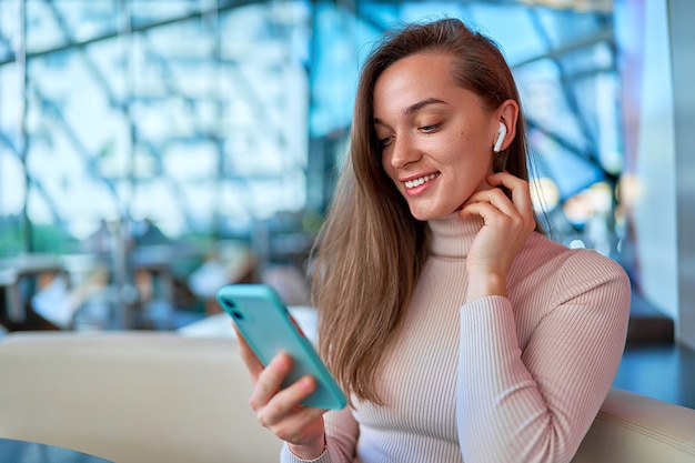 Jovem, moderna, casual, feliz e alegre, usando fones de ouvido sem fio, usando o smartphone para assistir a vídeos e ler notícias online enquanto está sentado e descansando em um café