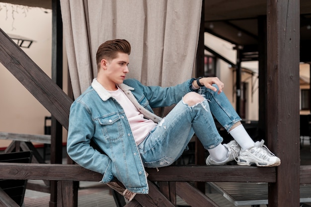 Jovem moderno com roupas da moda jeans azul casuais em tênis elegantes relaxa no corrimão de madeira na cidade. Cara de moda atraente em jeans vintage jovem usa ao ar livre. Moda masculina de verão.