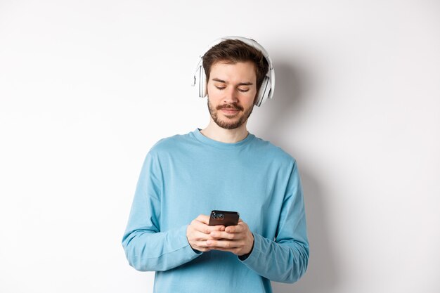 Jovem moderno de moletom azul coloca música no smartphone, ouvindo músicas em fones de ouvido sem fio, fundo branco