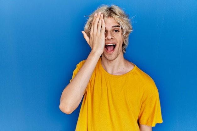 Jovem moderno de pé sobre fundo azul cobrindo um olho com a mão, sorriso confiante no rosto e emoção surpresa.