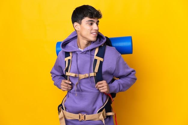 Foto jovem montanhista com uma grande mochila sobre amarelo isolado, olhando para o lado e sorrindo