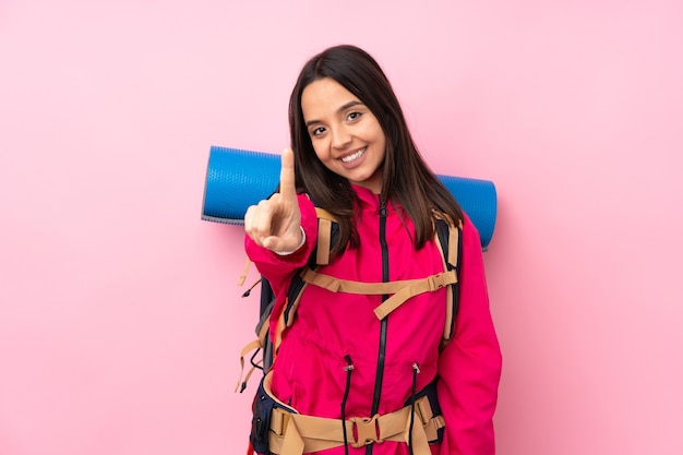 Jovem montanhista com uma grande mochila sobre uma parede rosa isolada, mostrando e levantando um dedo