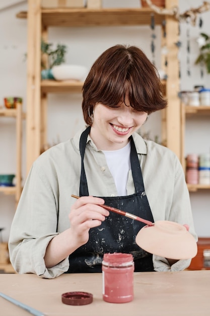 Jovem morena alegre em roupas de trabalho pintando tigela de barro artesanal
